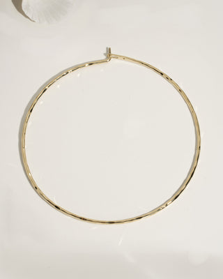 Single large gold hammered thin hoop earring laid flat on cream surface